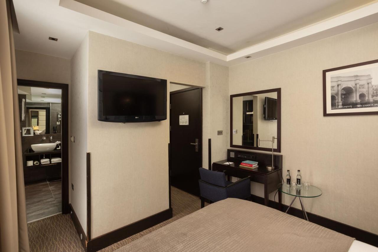 Montcalm Brewery Townhouse Hotel Londres Exterior foto The photo shows a hotel room featuring a double bed with a neutral-colored bedspread. On one wall, there is a flat-screen TV mounted. Adjacent to the bed, there is a stylish desk with a chair and a small glass table. A mirror is positioned above the 