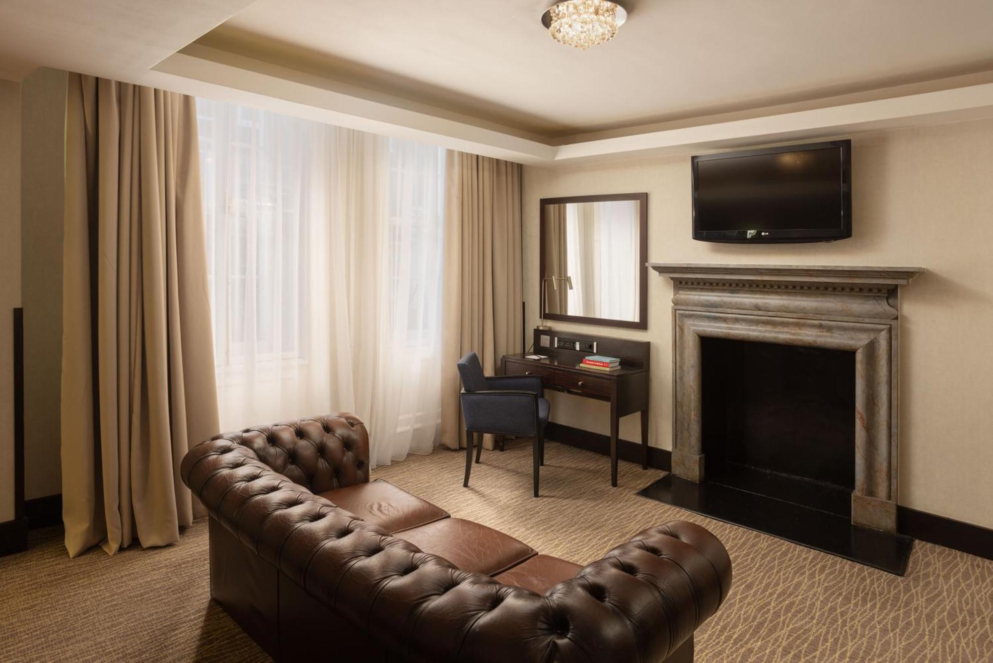 Montcalm Brewery Townhouse Hotel Londres Exterior foto The photo shows a stylish interior of a room, likely a living area or a hotel suite. It features a dark leather chesterfield sofa with tufted upholstery. Opposite the sofa is a small wooden desk with a chair, positioned near a wall-mounted flat-scree