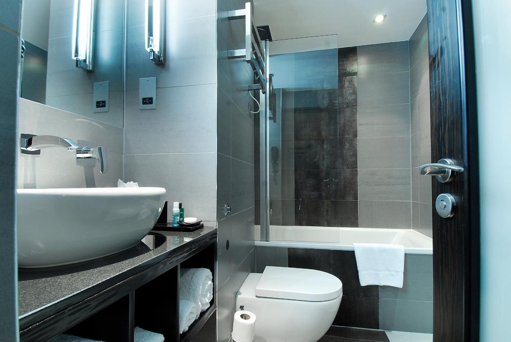 Montcalm Brewery Townhouse Hotel Londres Exterior foto The photo shows a modern bathroom with sleek design elements. It features a curved white sink on a dark countertop, with a mirror above that is illuminated by stylish lighting fixtures. Next to the sink, there's a toilet, and on the opposite side, a 