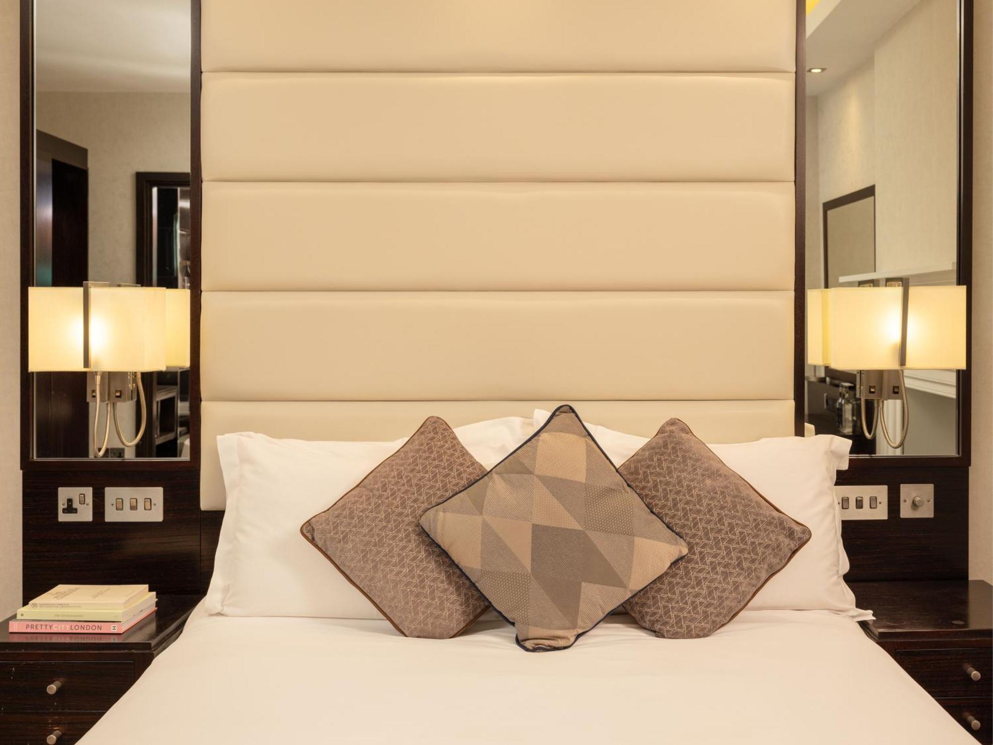 Montcalm Brewery Townhouse Hotel Londres Exterior foto The image shows a cozy bedroom setting. The bed features a headboard upholstered in a light beige fabric. It is adorned with three decorative pillows in varying patterns and colors, placed on white bedding. On either side of the bed, there are modern