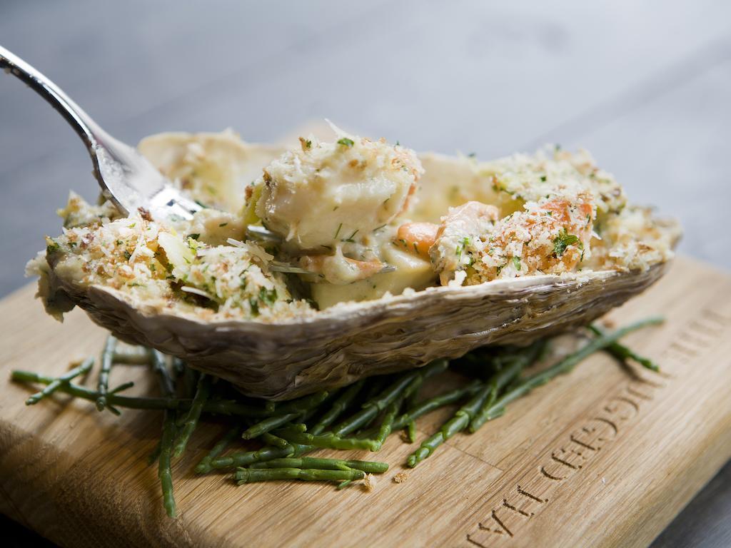 Montcalm Brewery Townhouse Hotel Londres Exterior foto The photo shows a large oyster shell filled with a mixture that appears to include seafood, herbs, and breadcrumbs. The dish is presented on a wooden board with some greenery, possibly a type of seaweed or garnishing. A fork is placed beside the oyst