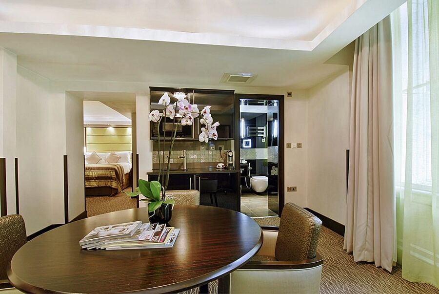 Montcalm Brewery Townhouse Hotel Londres Exterior foto The photo shows a stylish hotel room or suite. In the foreground, there is a round dining table with a few magazines on it. Surrounding the table are upholstered chairs. To the right, there are large windows with light-colored curtains, allowing natu