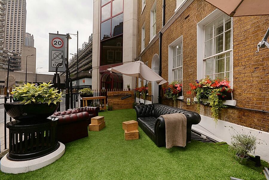 Montcalm Brewery Townhouse Hotel Londres Exterior foto The photo depicts a charming outdoor space, likely a patio or garden area. It features two comfortable-looking leather sofas, one of which is a deep burgundy color, placed on a green artificial lawn. There are wooden crates that could serve as side t